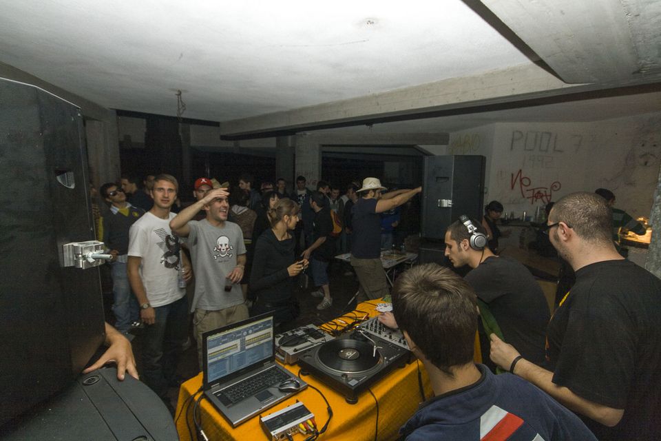Crowd around DJ decks on an orange cloth in a concrete room