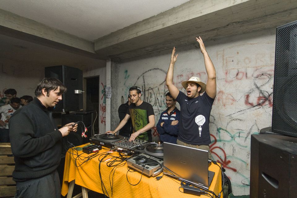 DJs at turntables by a graffiti wall as a dancer raises his arms