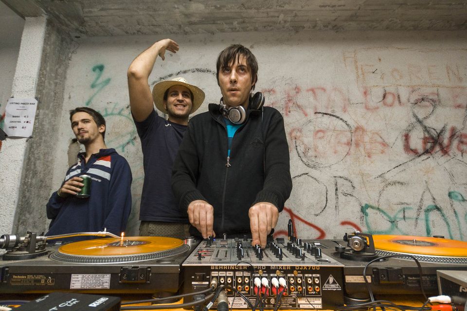 DJs behind turntables and a mixer in front of a graffiti wall