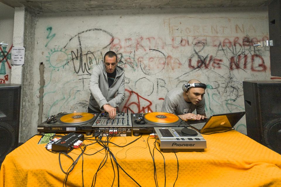 Two DJs at turntables on an orange cloth before a graffiti wall