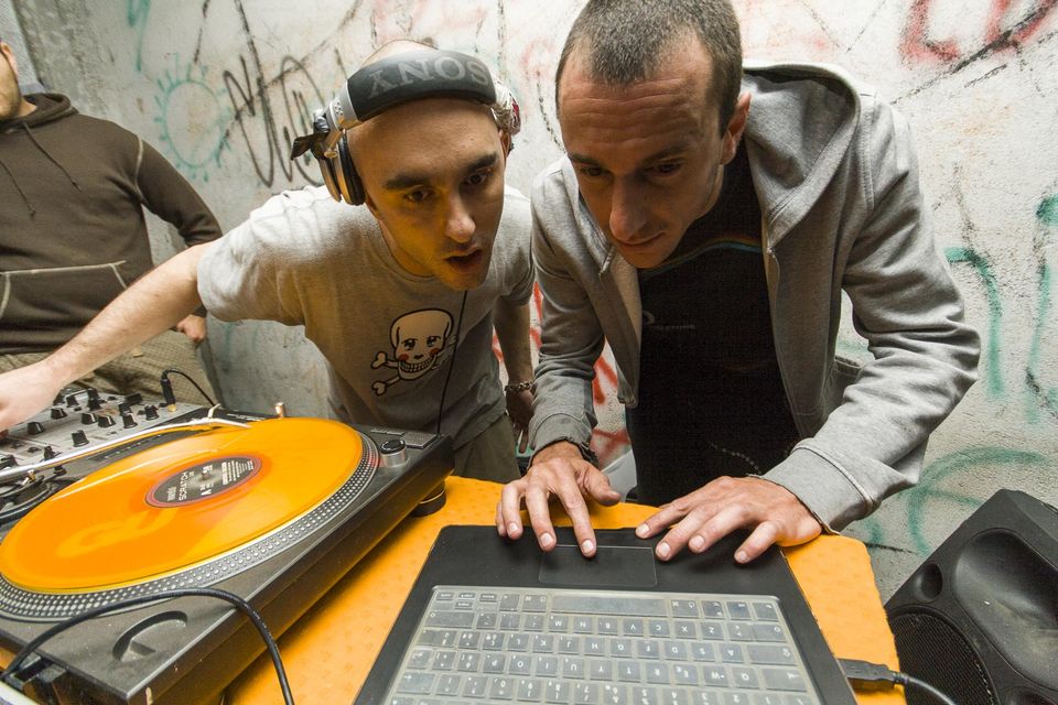 DJs bent over a laptop beside an orange turntable