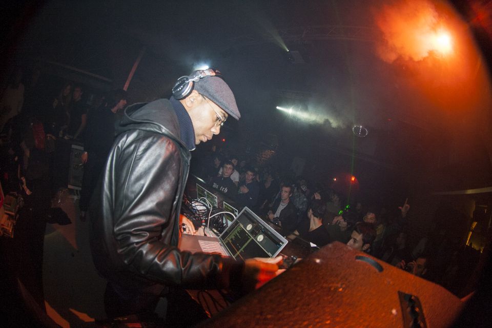 DJ in a flat cap, headphones and leather jacket bent over a laptop