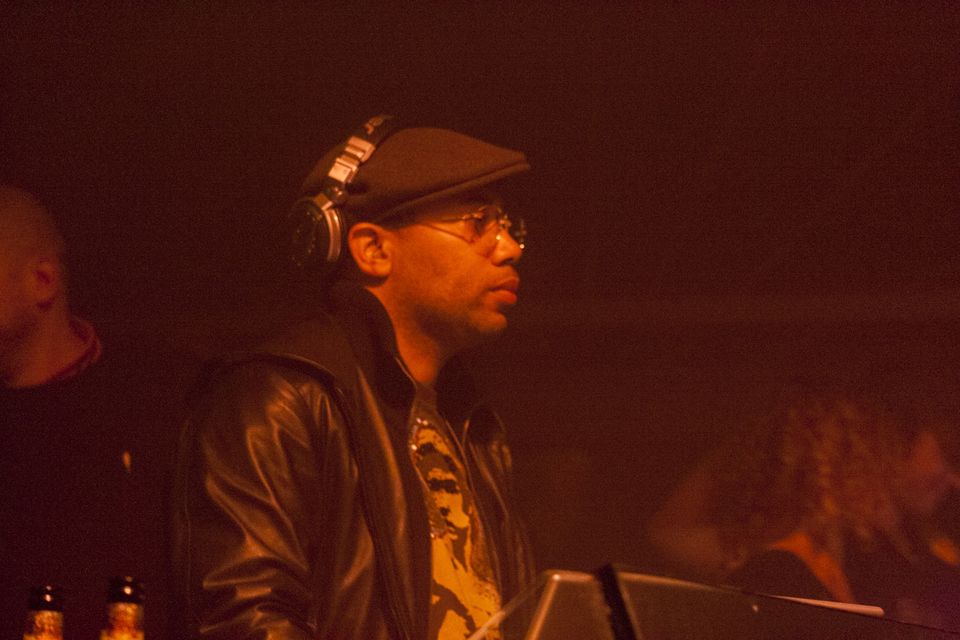 DJ in a flat cap and leather jacket standing in dim orange light