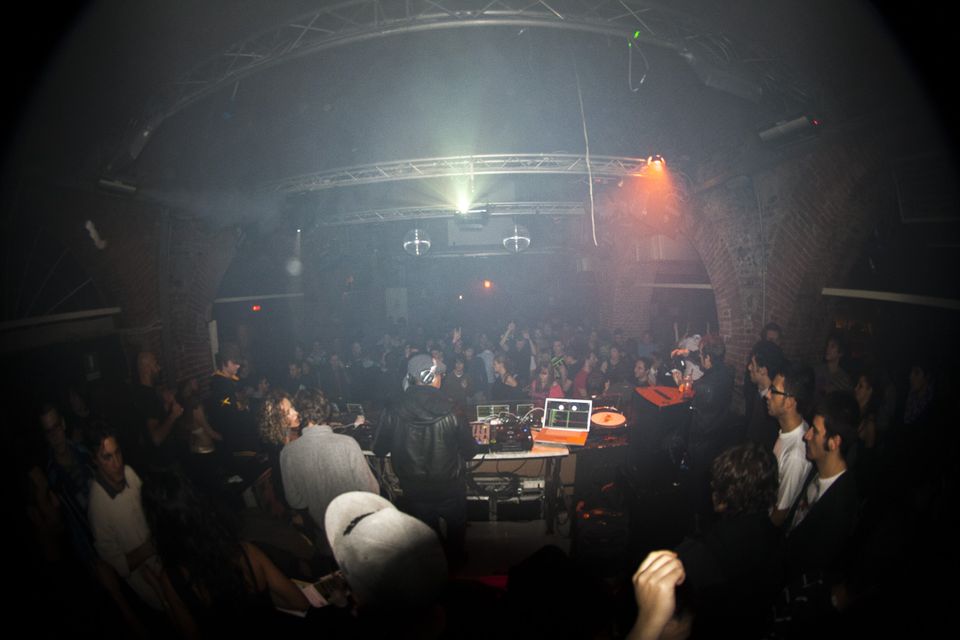 Smoky club with a crowd around the DJ booth under a steel truss
