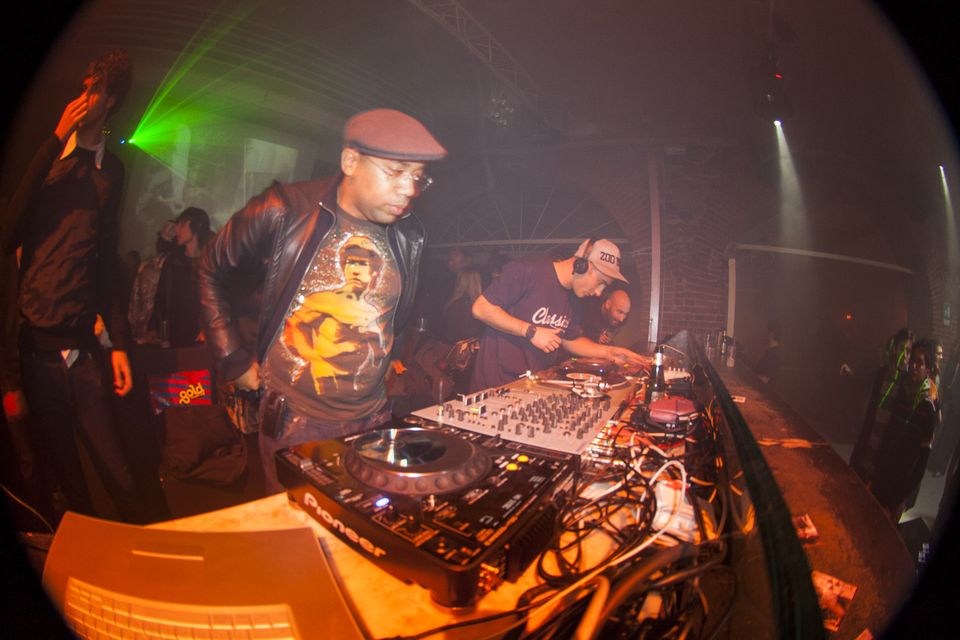 DJ in a flat cap and graphic T-shirt beside another at the mixer