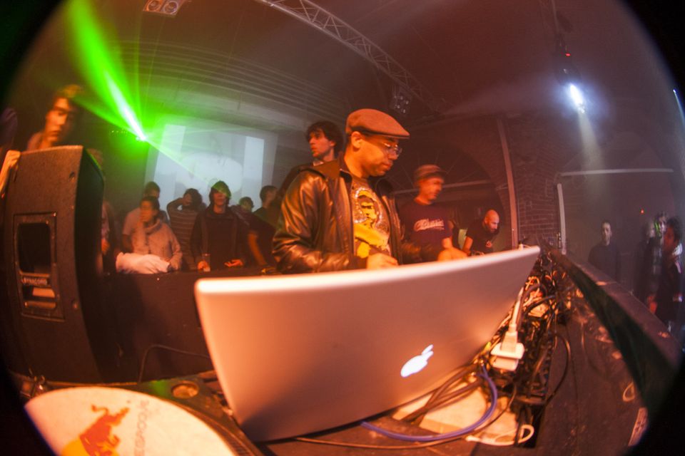 DJ in a flat cap behind a laptop with a slipmat in the foreground
