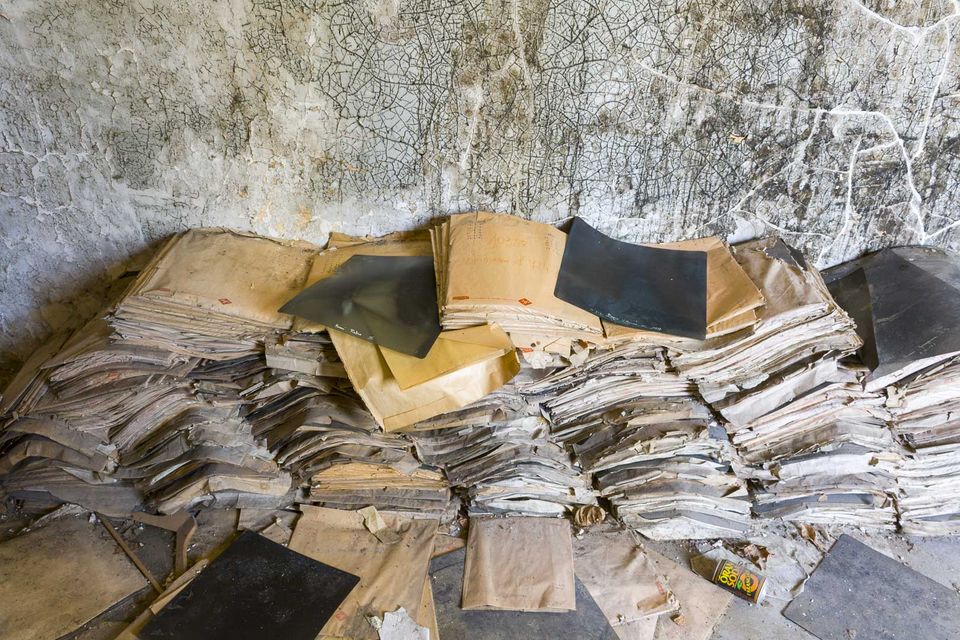 Stacks of yellowed paper records piled against a cracked wall