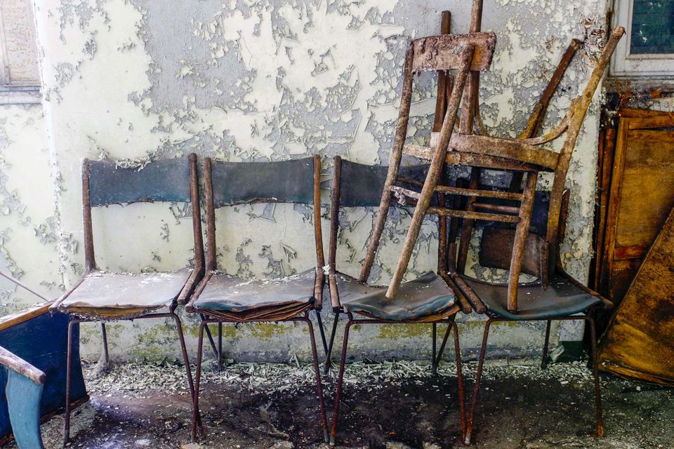 Four worn chairs with a broken one stacked against a flaking wall