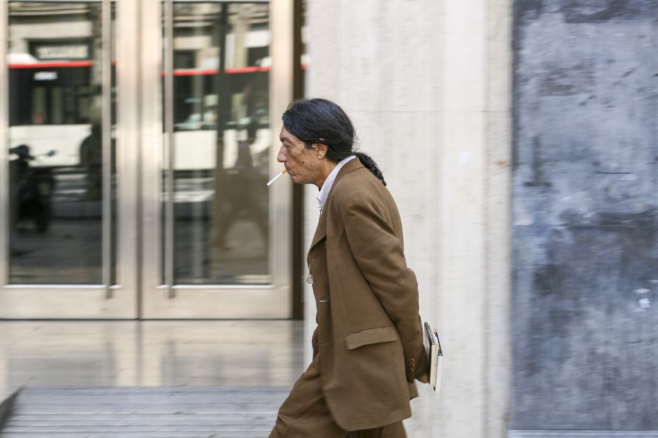Man with a cigarette in a brown coat walking past glass doors