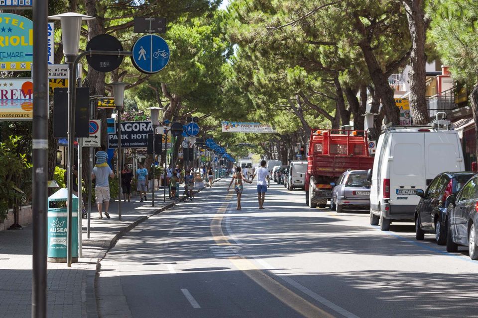 Shopping street lined with umbrella pines, pedestrians and parked vans