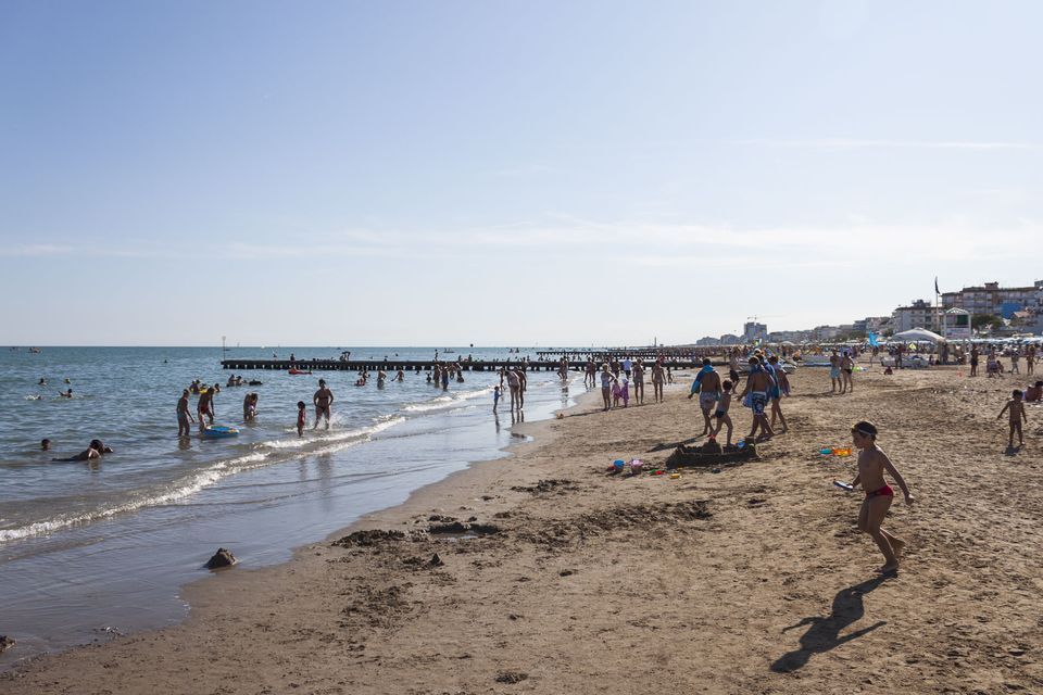 Crowded beach with swimmers in the shallows and a breakwater beyond