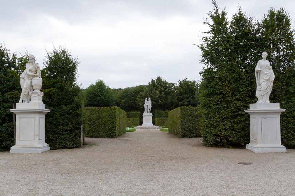 White statues on pedestals either side of a gravel path between hedges