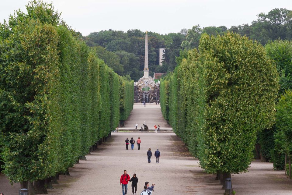 Avenue of clipped hedges leading to an obelisk and fountain with walkers