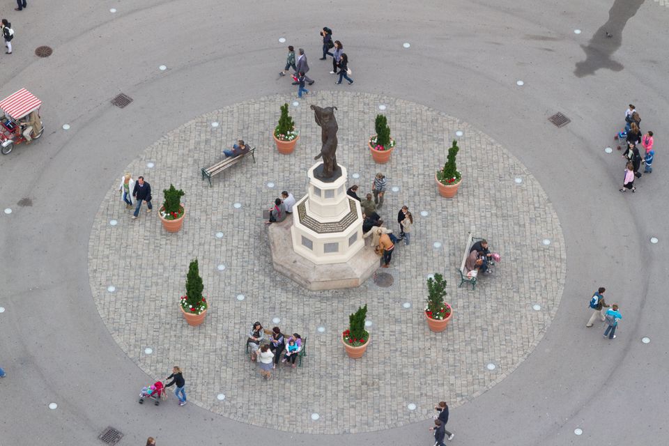 Circular cobbled square seen from above with fountain statue and potted trees