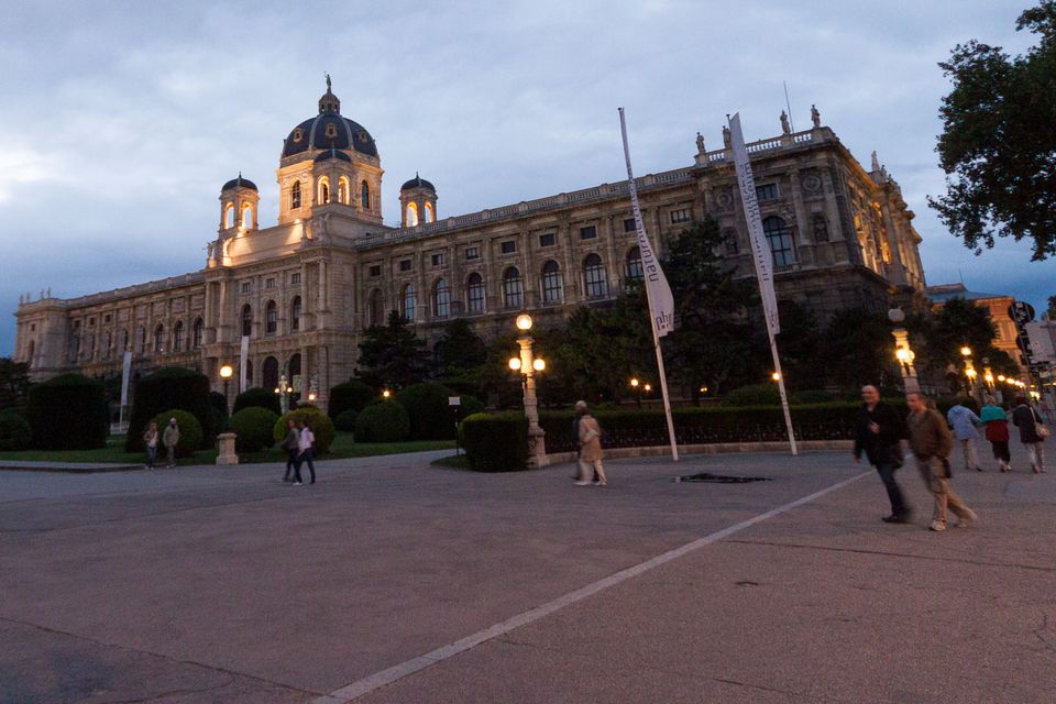 Domed museum facade lit at dusk with people crossing the square