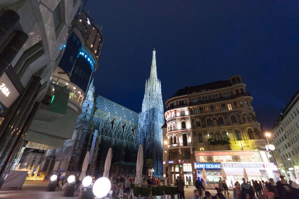 Gothic cathedral spire at night above café terraces and shop lights