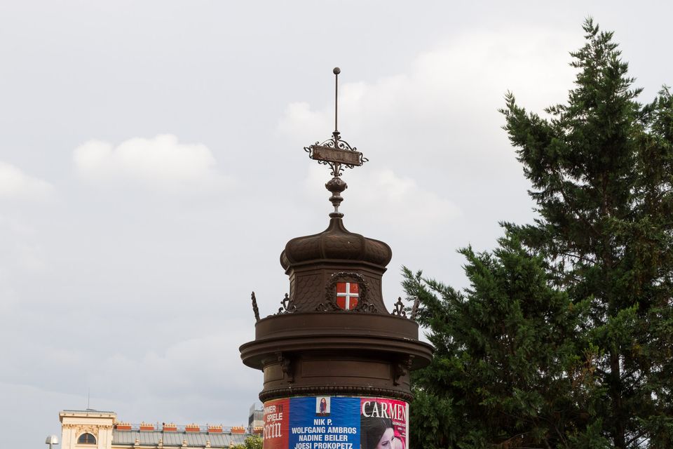 Dark ironwork kiosk with a red cross shield above a Carmen poster