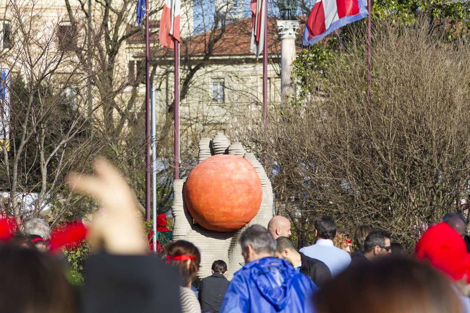 Large orange sculpture held in stone hands above the crowd